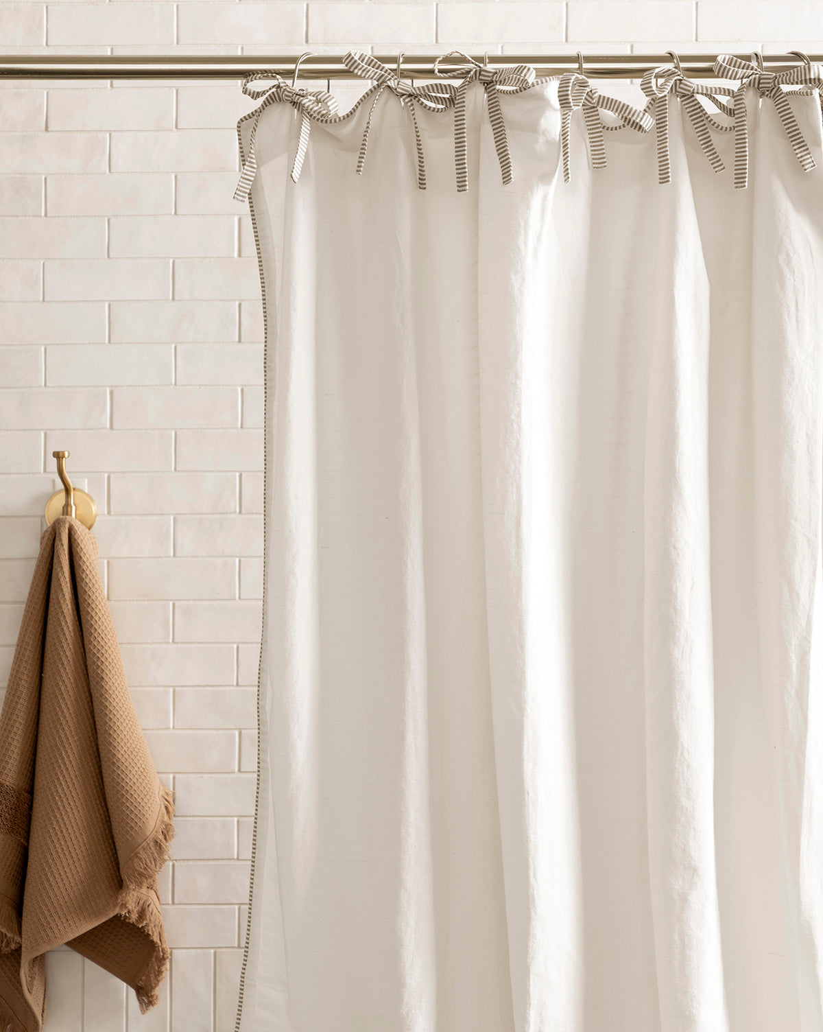 The Lillian Shower Curtain with Striped Ties hangs from a rod in front of light beige tiles, while a brown textured towel is displayed on a gold hook to the left, by McGee & Co.