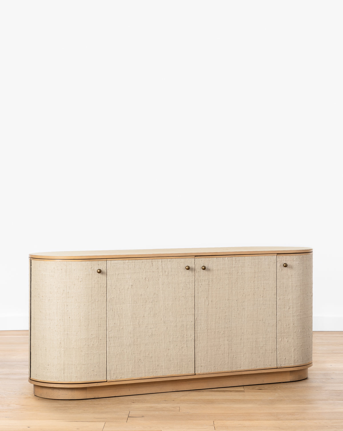 The Linu Woven Sideboard features rounded edges and textured beige fabric with four doors and small round knobs, set on a light wooden floor against a plain white background by McGee & Co.