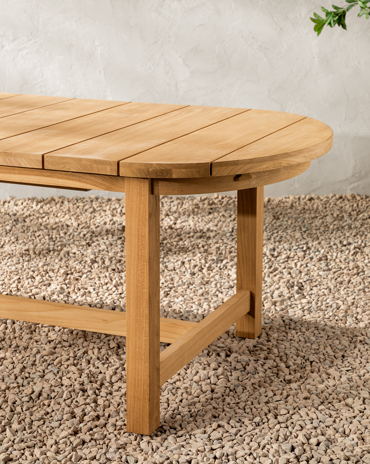 The Linwood Teak Extension Dining Table features a slatted oval top and sturdy legs, shown here in close-up on pebbles against a light textured wall, by McGee & Co.