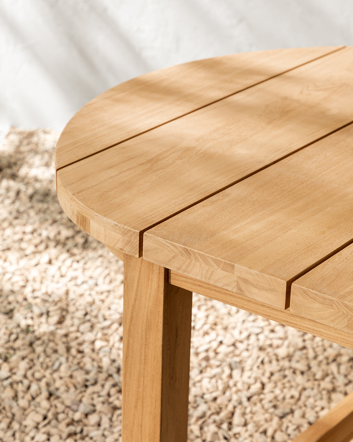 Close-up of the Linwood Teak Extension Dining Table with a slatted top, set on light-colored pebbles as sunlight casts soft shadows across the table and ground, by McGee & Co.