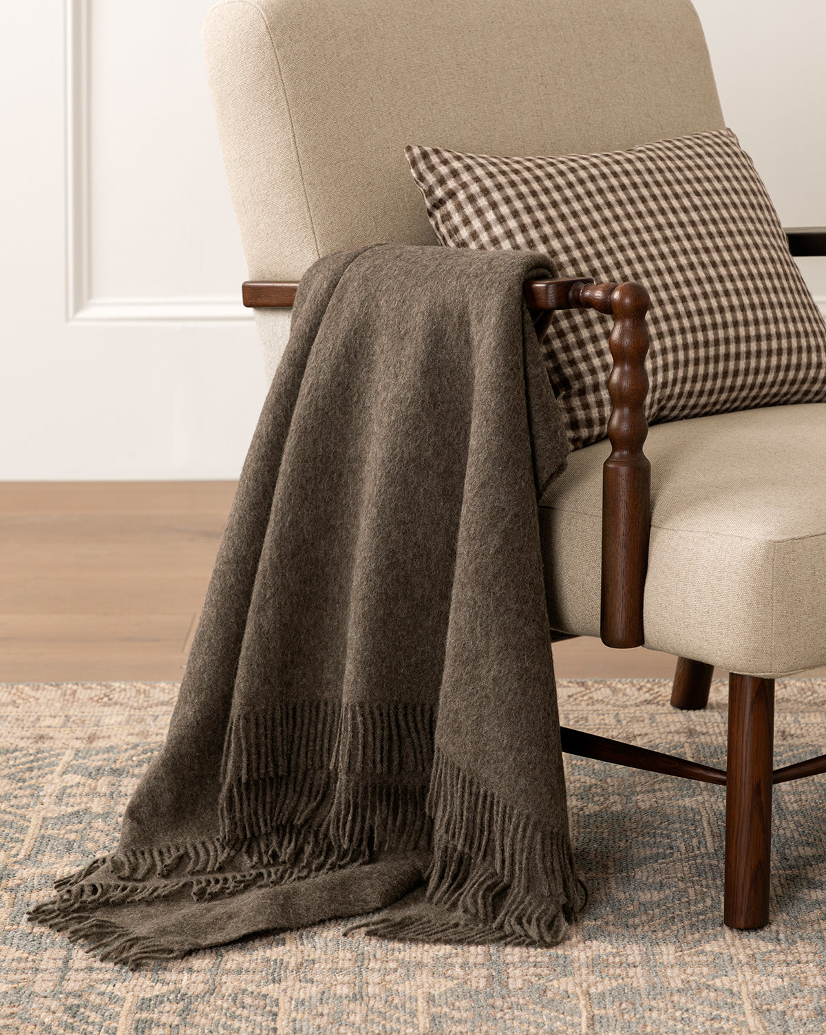The Fibre Locester Throw blanket is draped over a cozy lounge chair with a with delicate fringe at the ends from McGee & Co
