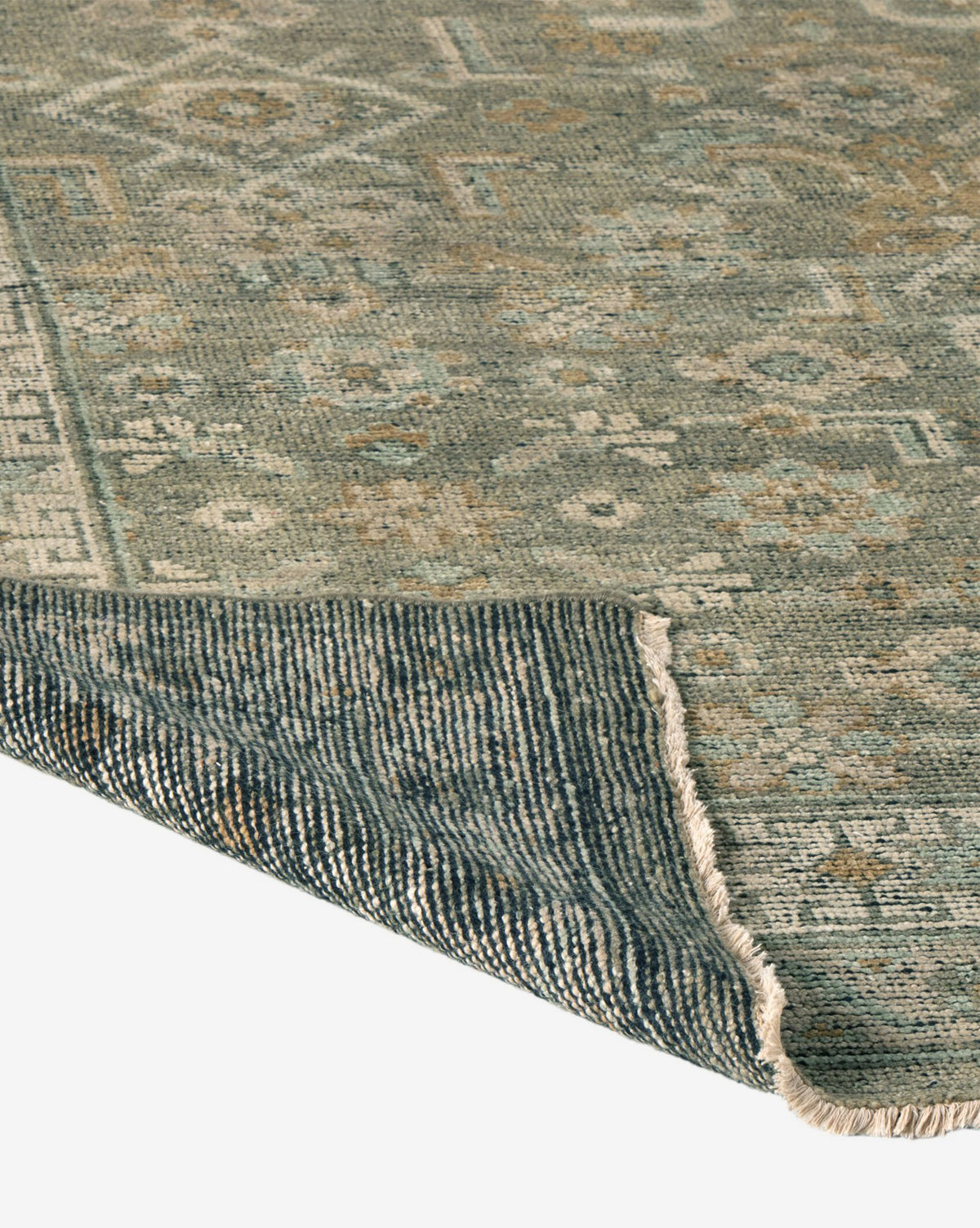 A close-up of the Loreo Hand-Knotted Wool Rug highlights its muted blue and beige geometric pattern, showcasing the detailed texture with a corner folded to reveal the underside, by McGee & Co.