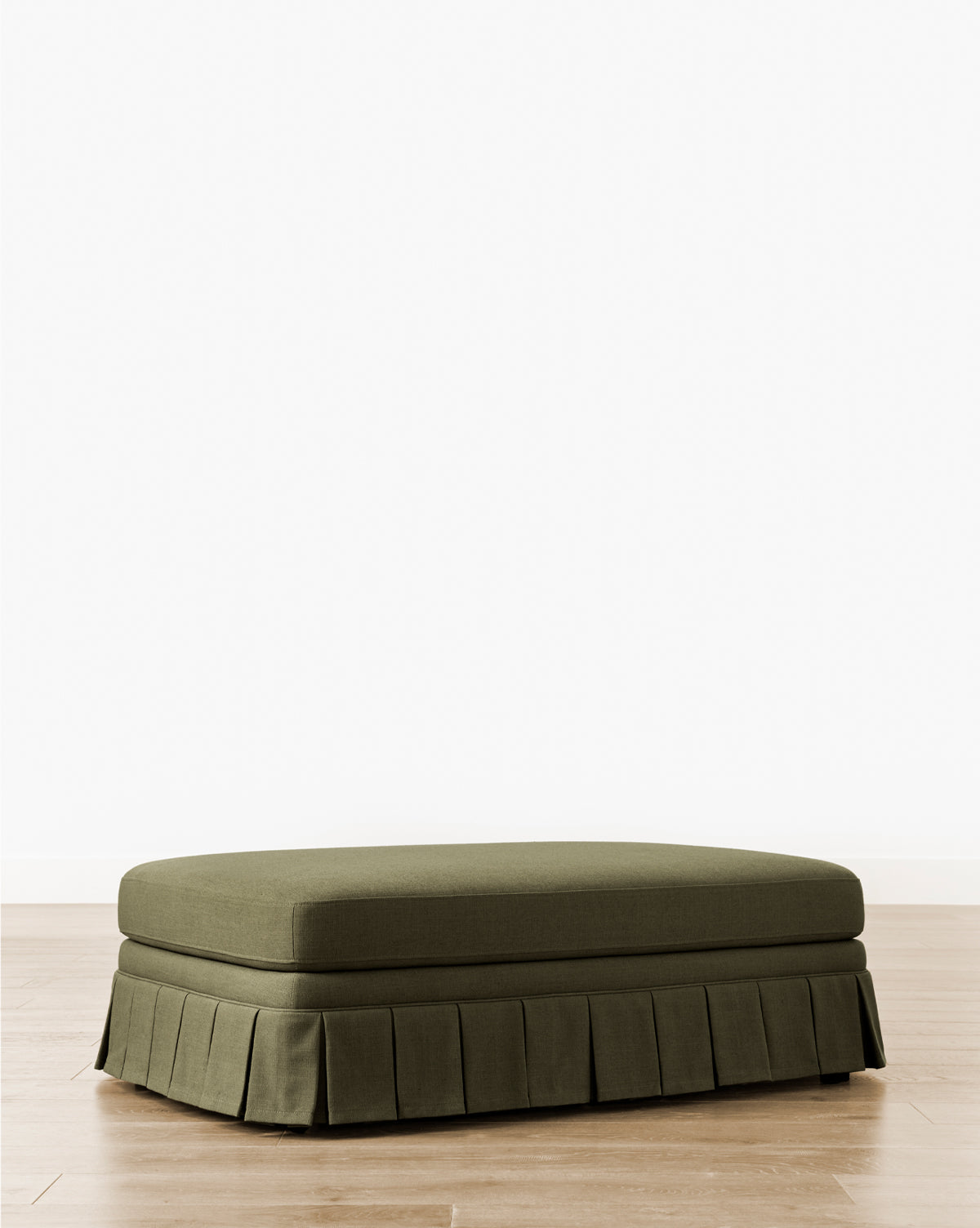 The Louisa Ottoman showcases custom olive green upholstery and a box pleat skirt, displayed elegantly on a light wood floor with a plain white background, by McGee & Co.