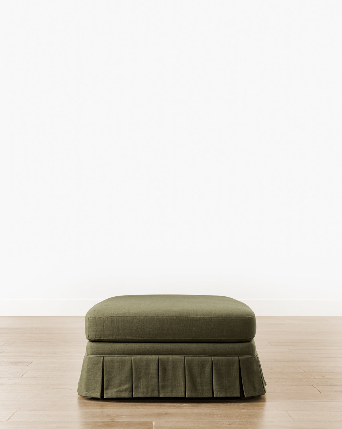 The Louisa Ottoman showcases custom green upholstery and a tailored box pleat skirt, elegantly placed on a light wood floor with a simple white wall backdrop, by McGee & Co.