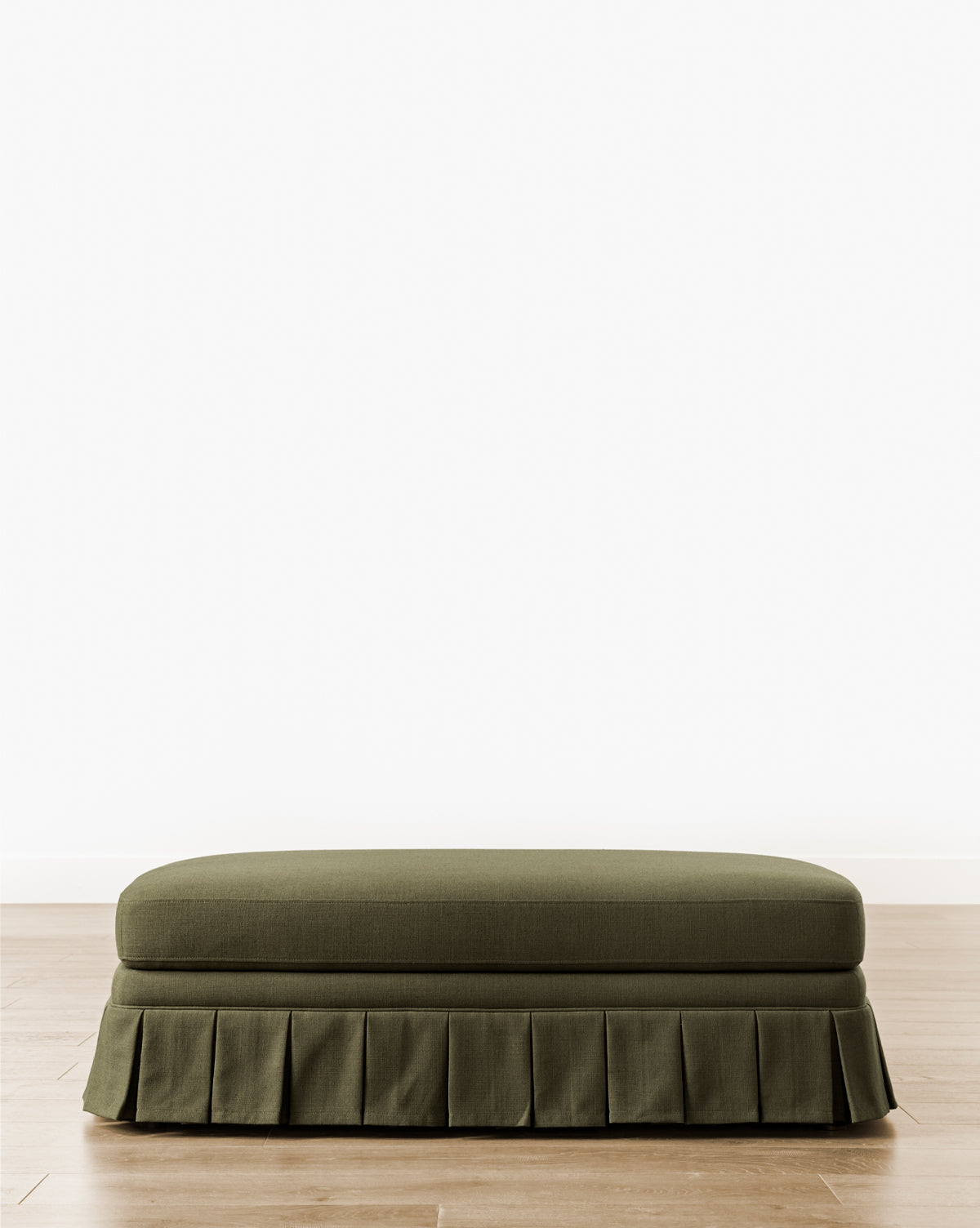 A Louisa Ottoman with green custom upholstery and a box pleat skirt rests on a light wood floor against a plain white wall, by McGee & Co.