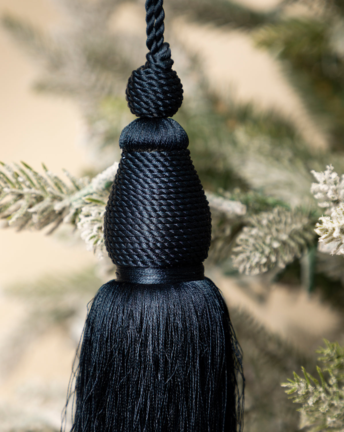 A close-up of the Lucia Bullion Tassel, a black vintage-inspired holiday ornament with silky threads, hanging on a snow-dusted artificial pine branch, by McGee & Co.