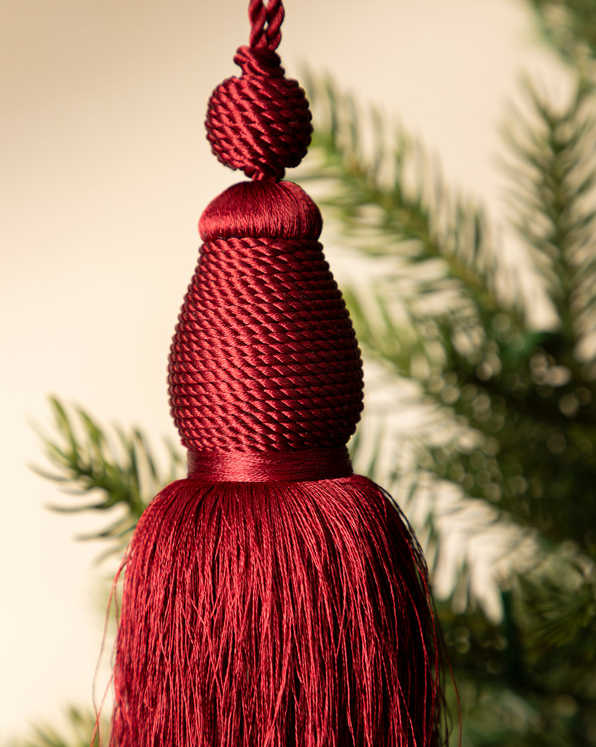 A close-up of the Lucia Bullion Tassel, a red holiday tree ornament, hanging with softly blurred green pine branches in the background, by McGee & Co.