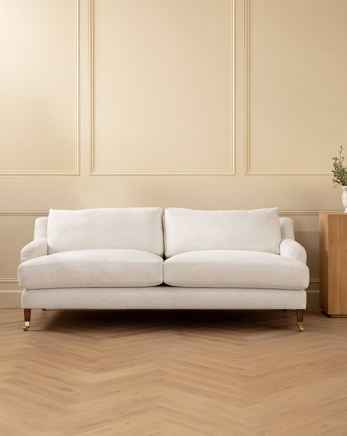 The Lucille English Roll Arm Sofa 90.5 in a modern cream hue, with plush down-wrapped cushions, sits before a beige paneled wall on light herringbone wood flooring. To the right, a small wooden side table holds a vase of greenery by McGee & Co.