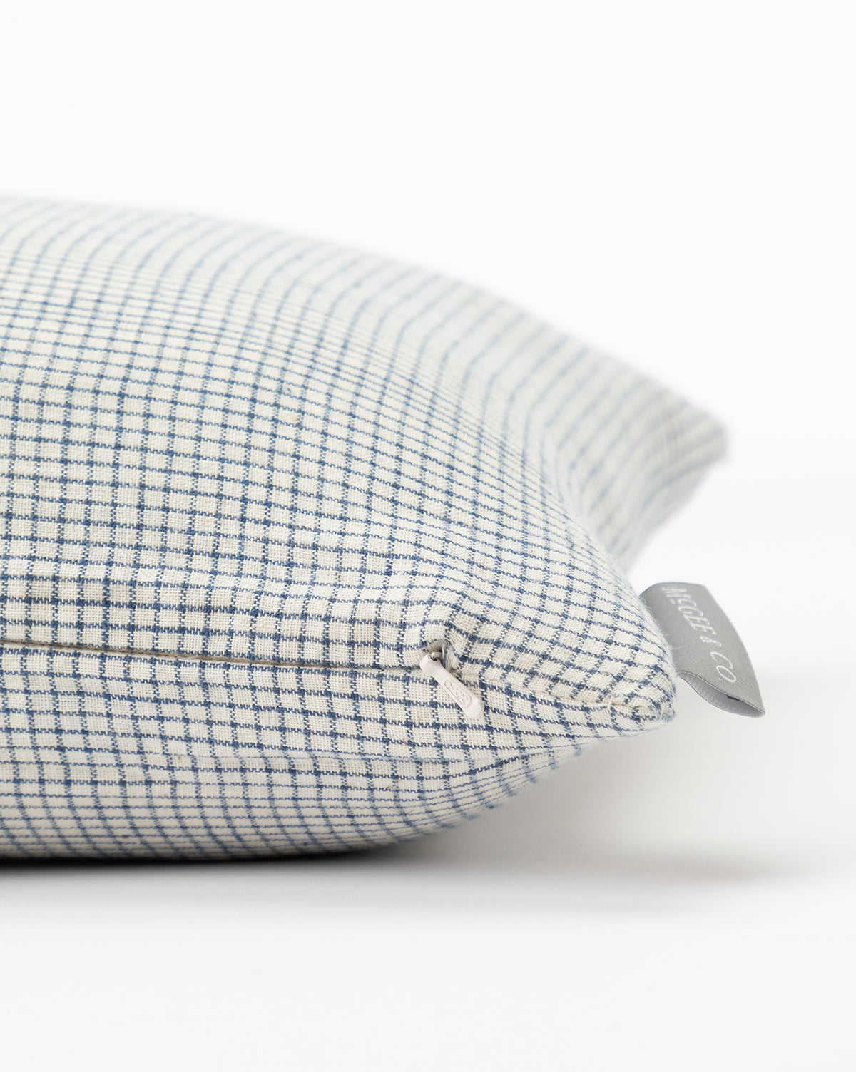 Close-up of the Luther Pillow Cover in blue and cream with a grid pattern and a gray fabric tag on the corner, shown resting on a white surface, by McGee & Co.