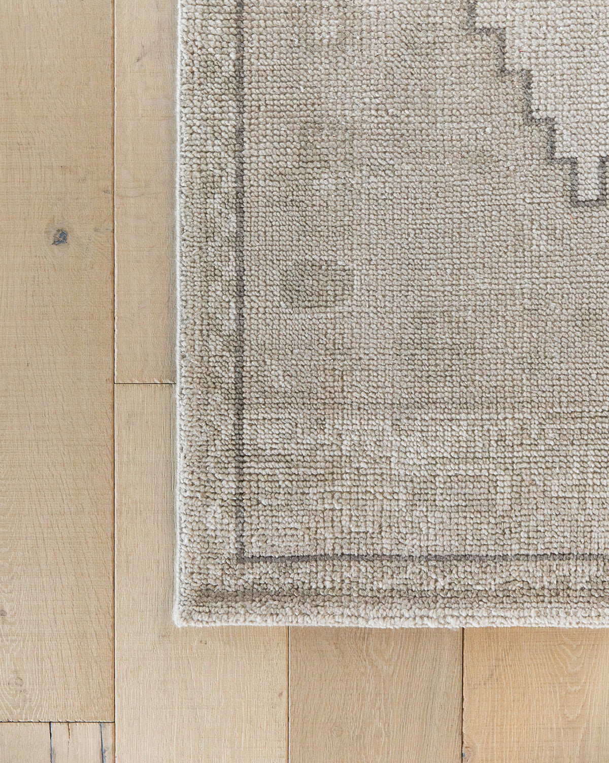 A close-up of the Pembroke Hand-Knotted Wool Rug, featuring a light tan textured transitional design with a subtle geometric border, shown on light wood flooring with its corner and part of the wooden floor visible, by McGee & Co.