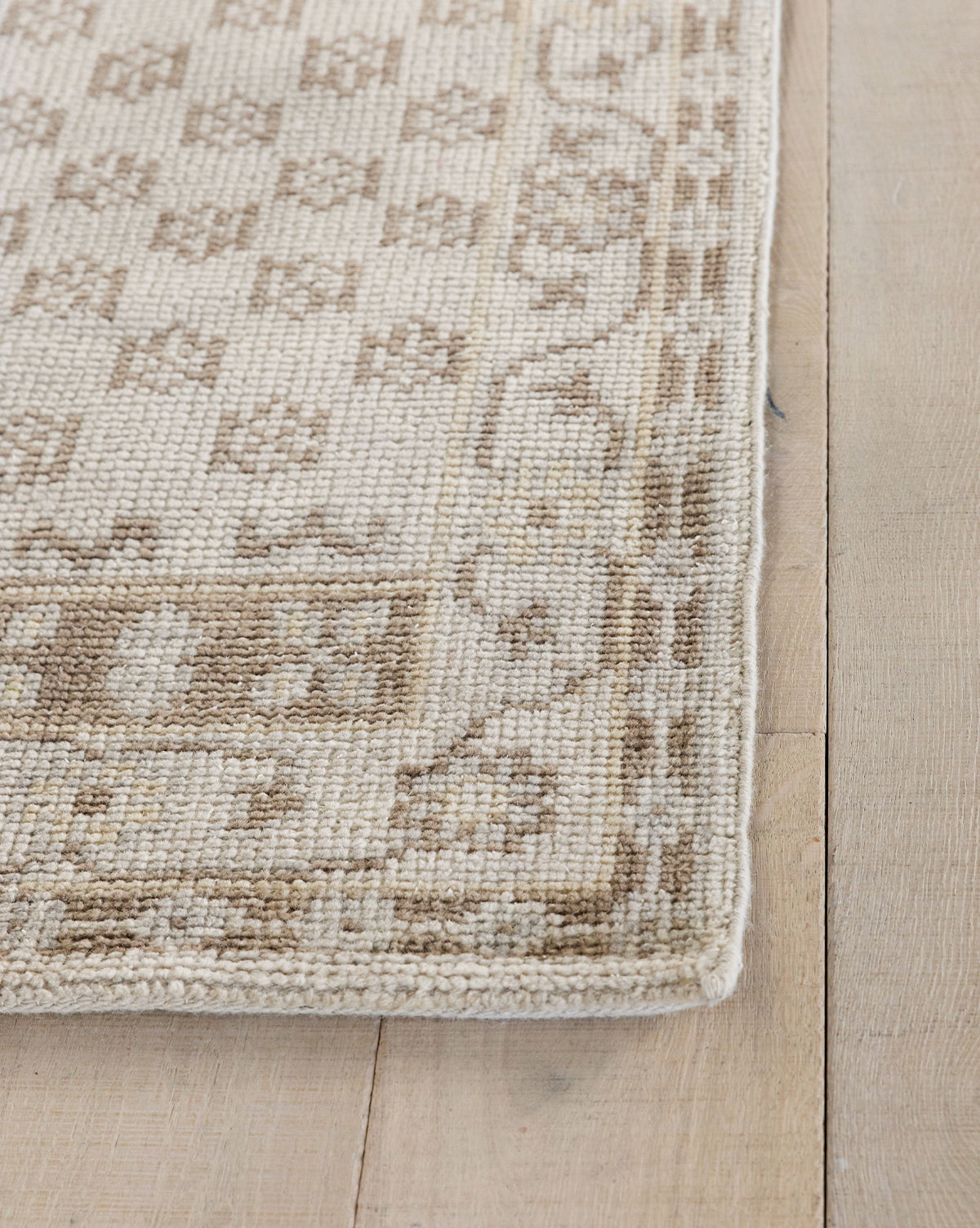 Close-up of the Anya Neutral Hand-Knotted Wool Rug in beige and light brown, featuring geometric and floral patterns. The detailed, durable woven texture is highlighted at the corner, displayed on a light wooden floor, by McGee & Co.
