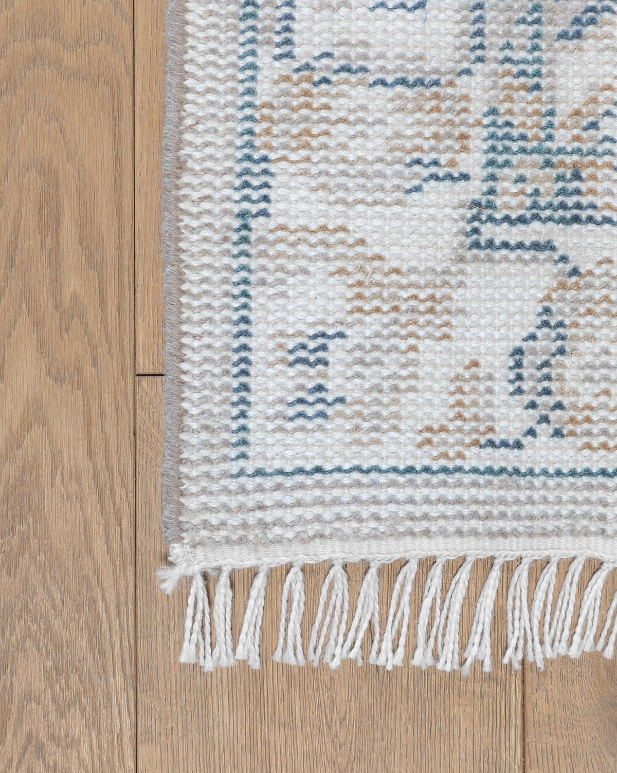 A close-up of the Merilyn Hand-Knotted Rug with white fringe, showcasing its subtle beige and blue pattern, displayed on a wooden floor, by McGee & Co.