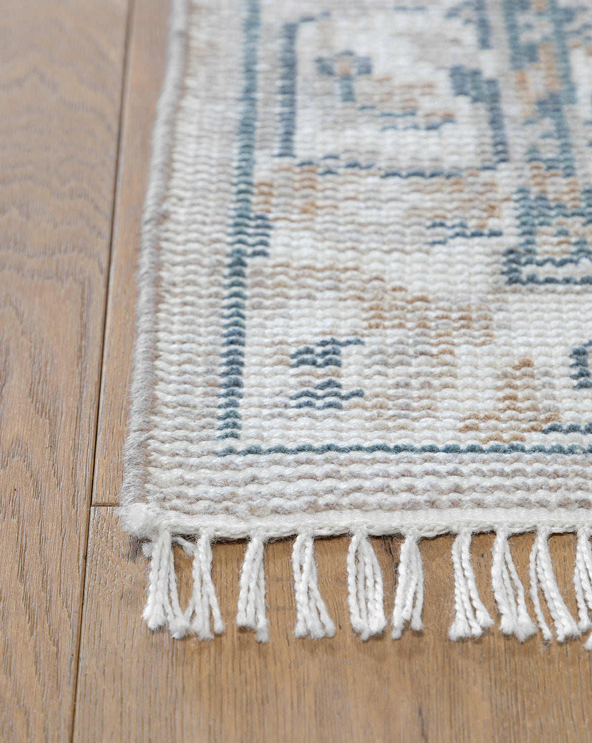 Close-up of the Merilyn Hand-Knotted Rug, featuring a beige and blue geometric pattern, white fringe accents, and fine craftsmanship. The rug is displayed elegantly on a light brown wooden floor, by McGee & Co.