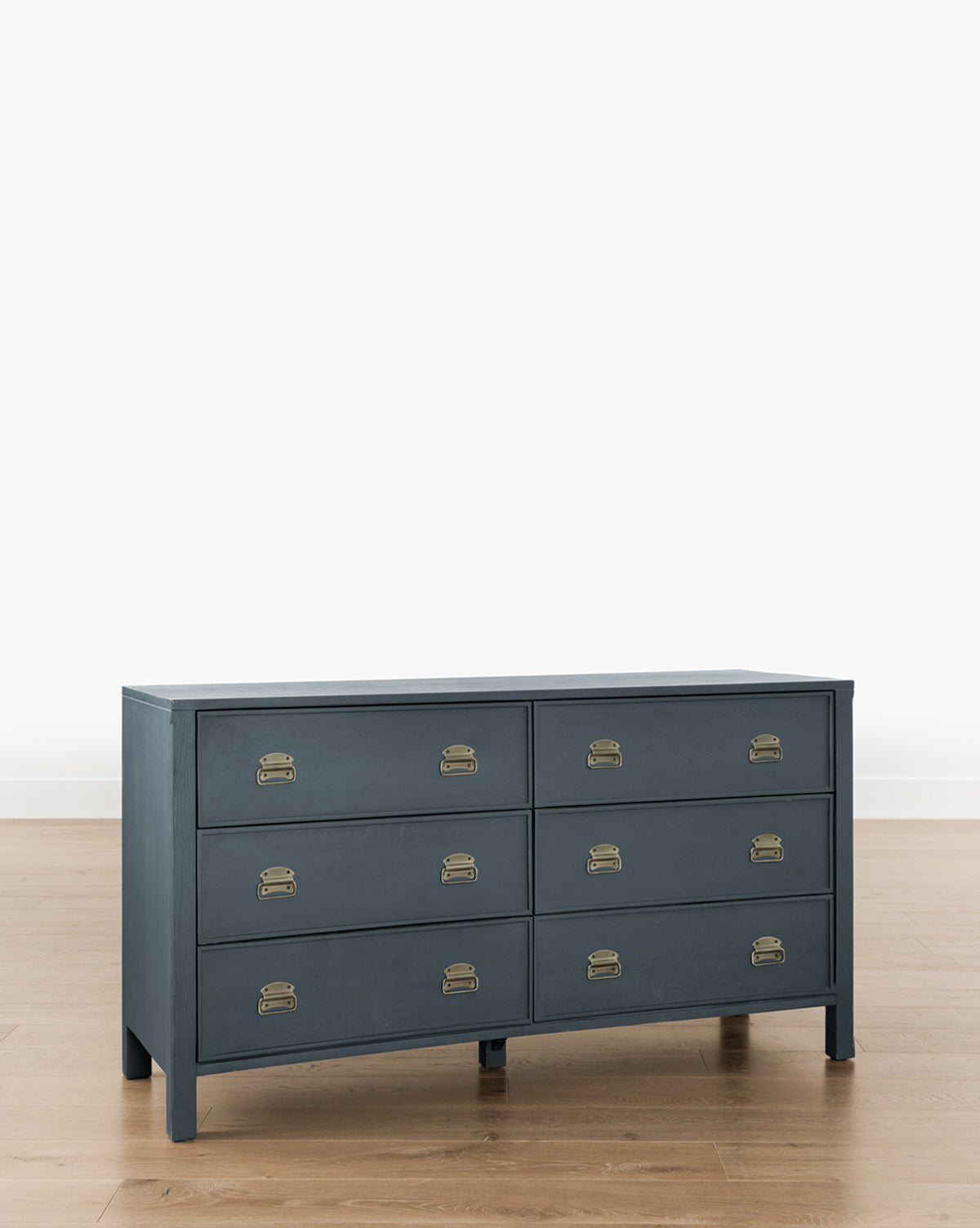 The Mackenna Dresser is a dark blue wooden dresser with six drawers and brass pulls, standing on a light wooden floor against a white wall.