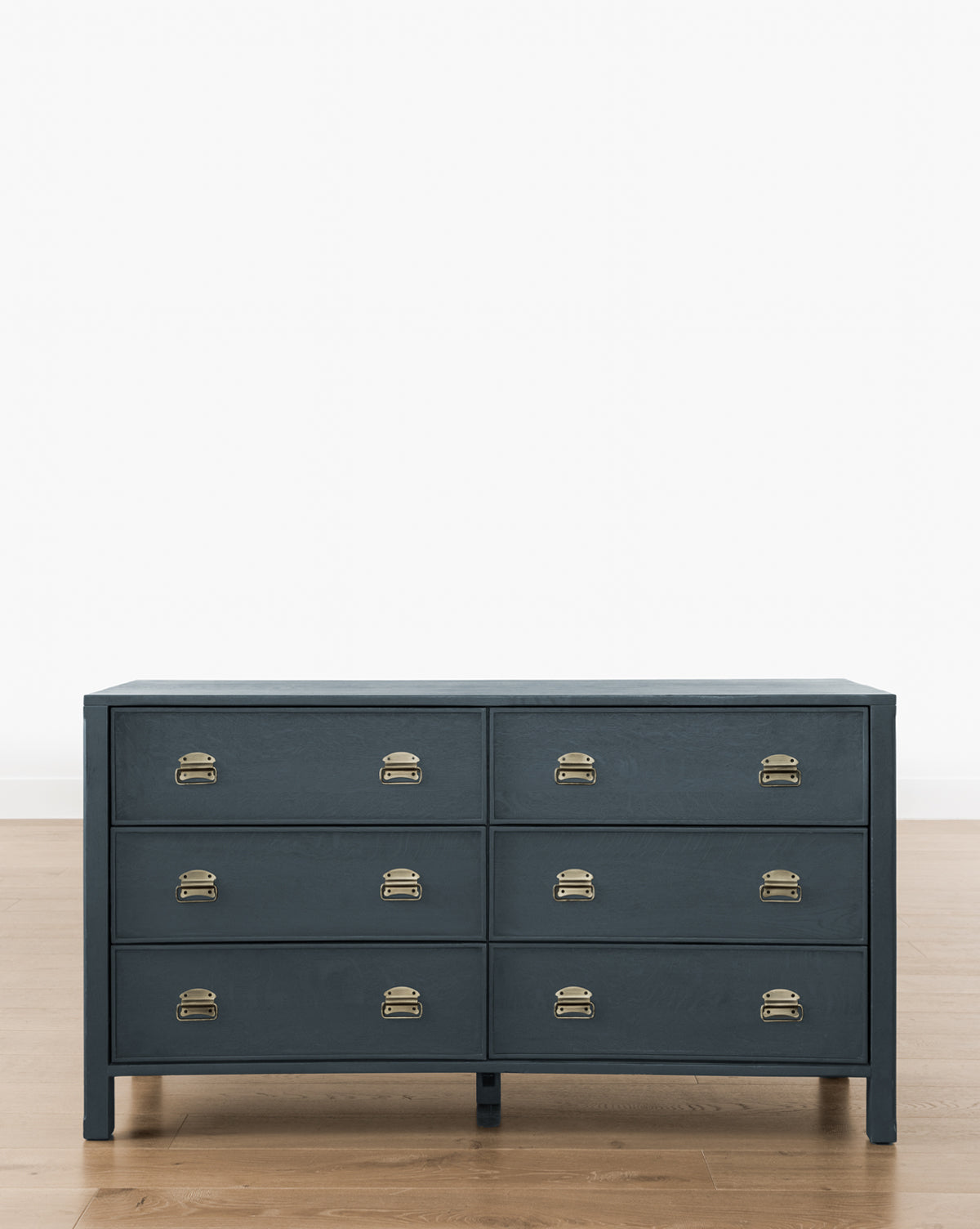 A Mackenna Dresser in dark blue, featuring six drawers with elegant brass pulls, stands on a wooden floor against a plain white wall.