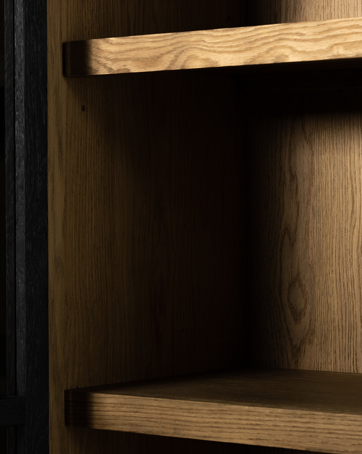 A close-up of three empty wooden shelves inside the Maddox Bookcase, showcasing natural wood grain and warm light highlighting the texture behind its elegant glass doors, by McGee & Co.