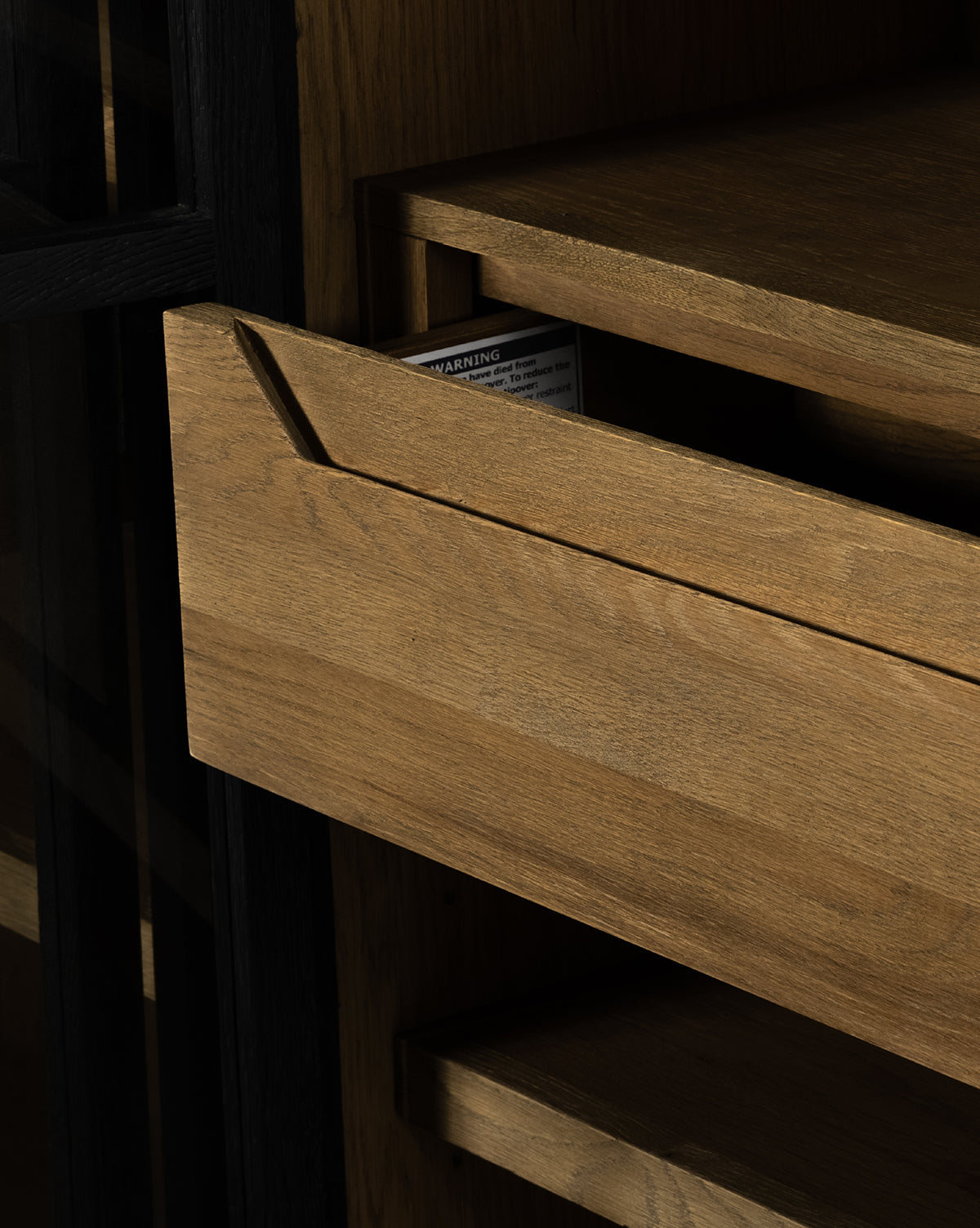 A partially opened drawer of the Maddox Bookcase reveals a warning label inside. The oak shelves and drawers are light brown, and a section of the black frame is visible on the left, by McGee & Co.