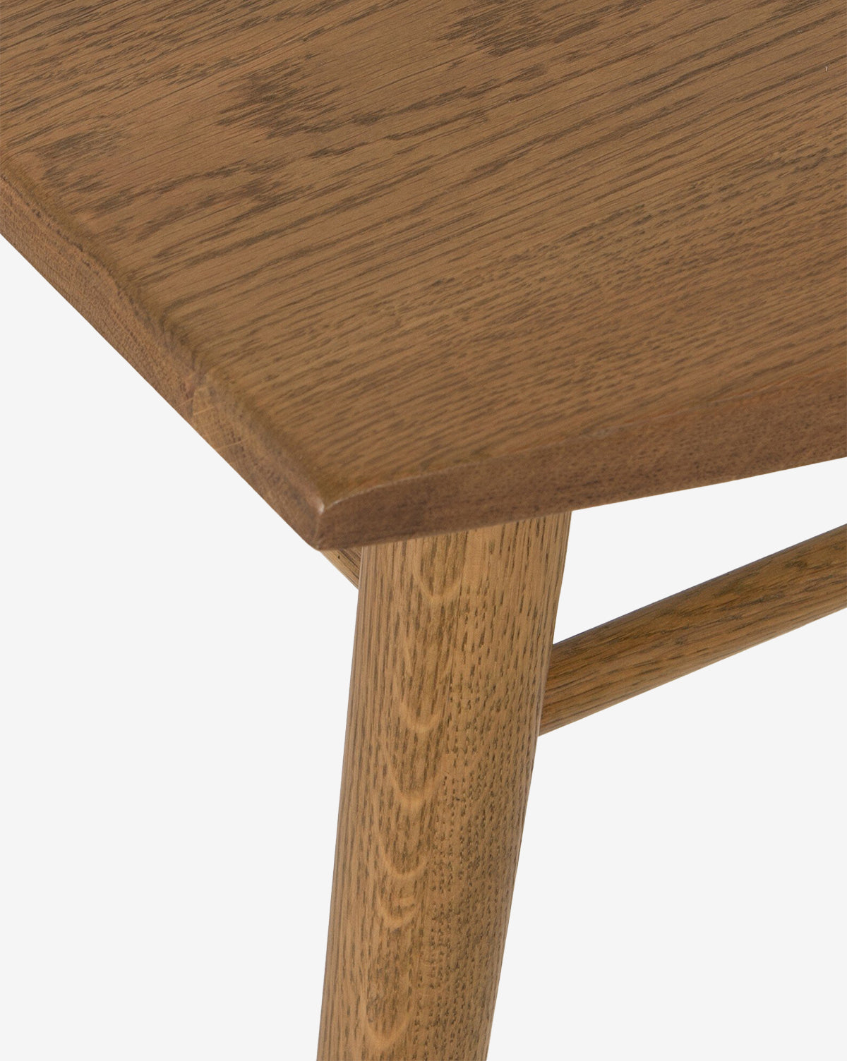 Close-up of a wooden table’s corner with smooth grain, showing part of the tabletop, leg, and support bar. The Madison Dining Chair is partially visible against a white background, by McGee & Co.