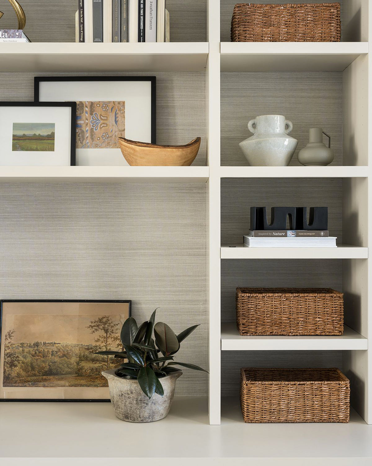 A white shelving unit displays framed art, woven baskets, the Malaga Vase with a white reactive glaze, a wooden bowl, and a potted plant—all neatly arranged for a stylish, modern look.