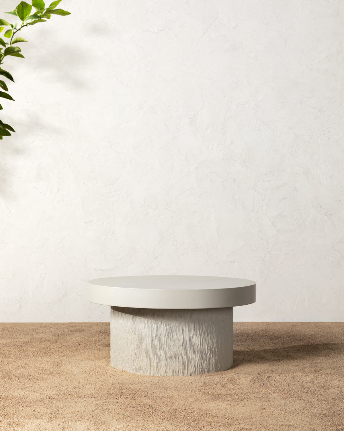 The Malcolm Fiberstone Coffee Table features a round, minimalist design with a textured cylindrical base and smooth top, ideal for refined outdoor decor. It sits on a sandy floor beside a light, textured wall with leafy greenery above by McGee & Co.