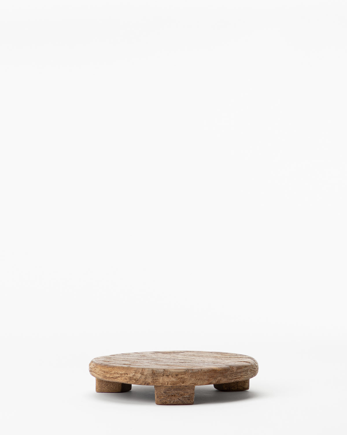 The Mango Wood Carved Pedestal is a small, round stand with four short legs, ideal as an elevated serving tray or rustic charcuterie board, shown against a plain white background.