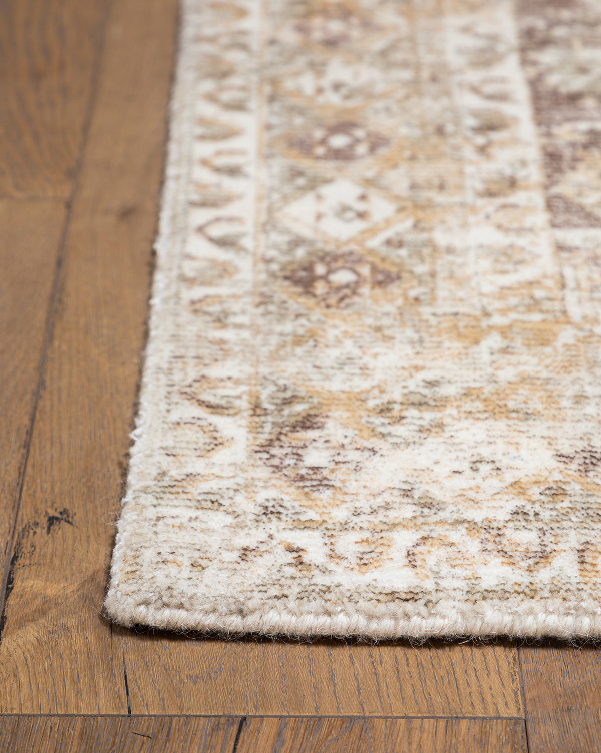 A close-up of the Marcella Handwoven Rug in neutral tan, beige, brown, and cream tones shows its subtle, faded geometric and floral pattern resting on a wooden floor, by McGee & Co.