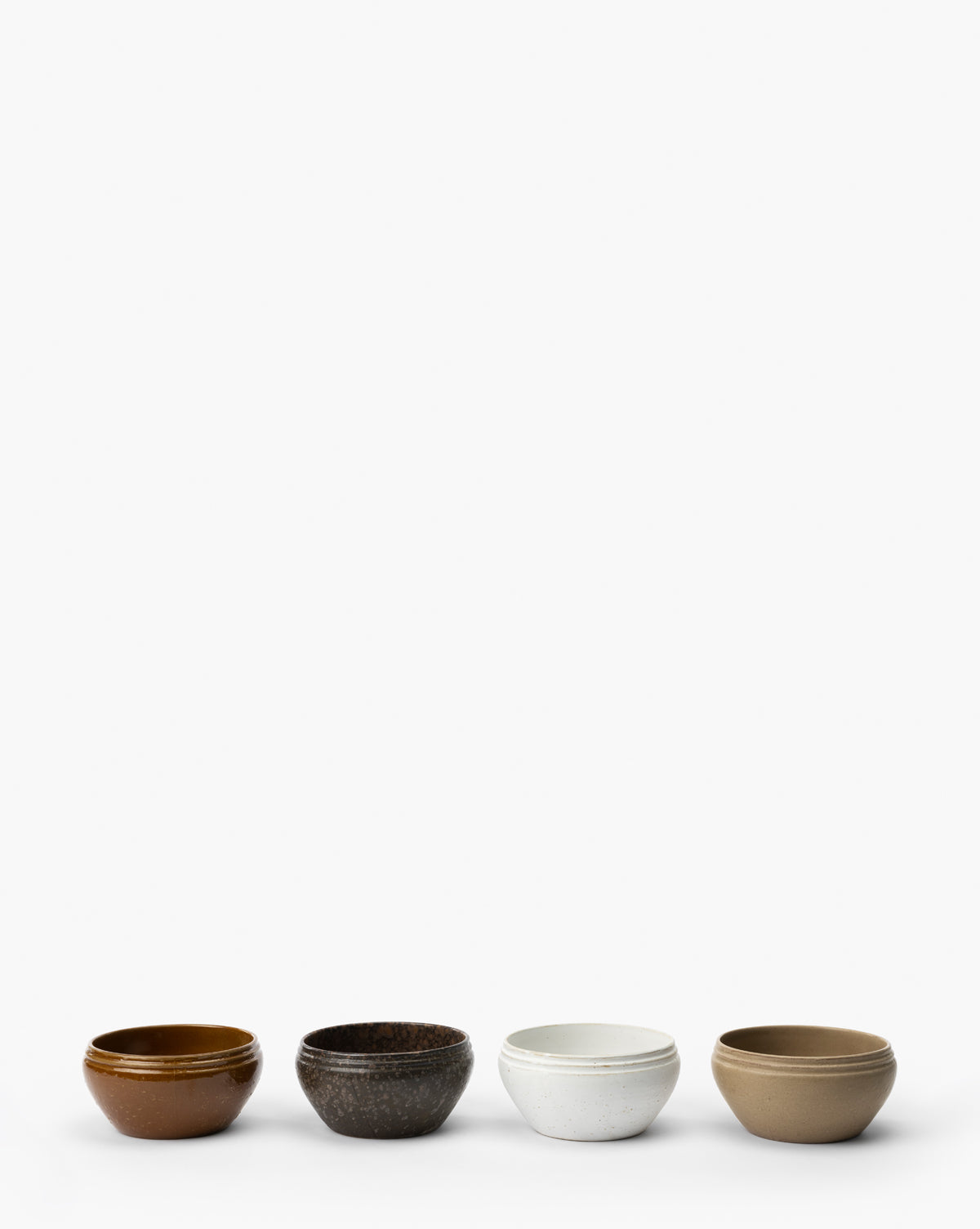 The Mariella Stoneware Bowls (Set of 4) features small, round bowls in brown, dark speckled, white speckled, and beige, displayed in a row on a white background to highlight their organic texture and handmade quality by McGee & Co.