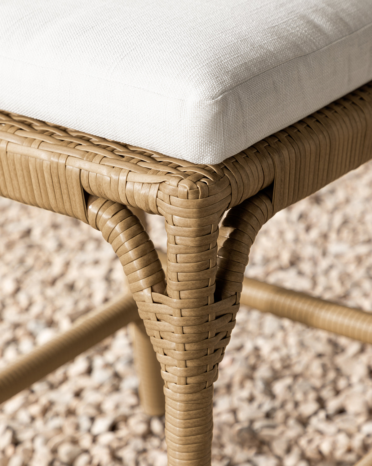 Close-up of the Makerspalm Marisol Indoor/Outdoor Counter Stool (Set of 2), showcasing a handwoven wicker leg and white cushioned seat, displayed on small beige pebbles. McGee & Co.