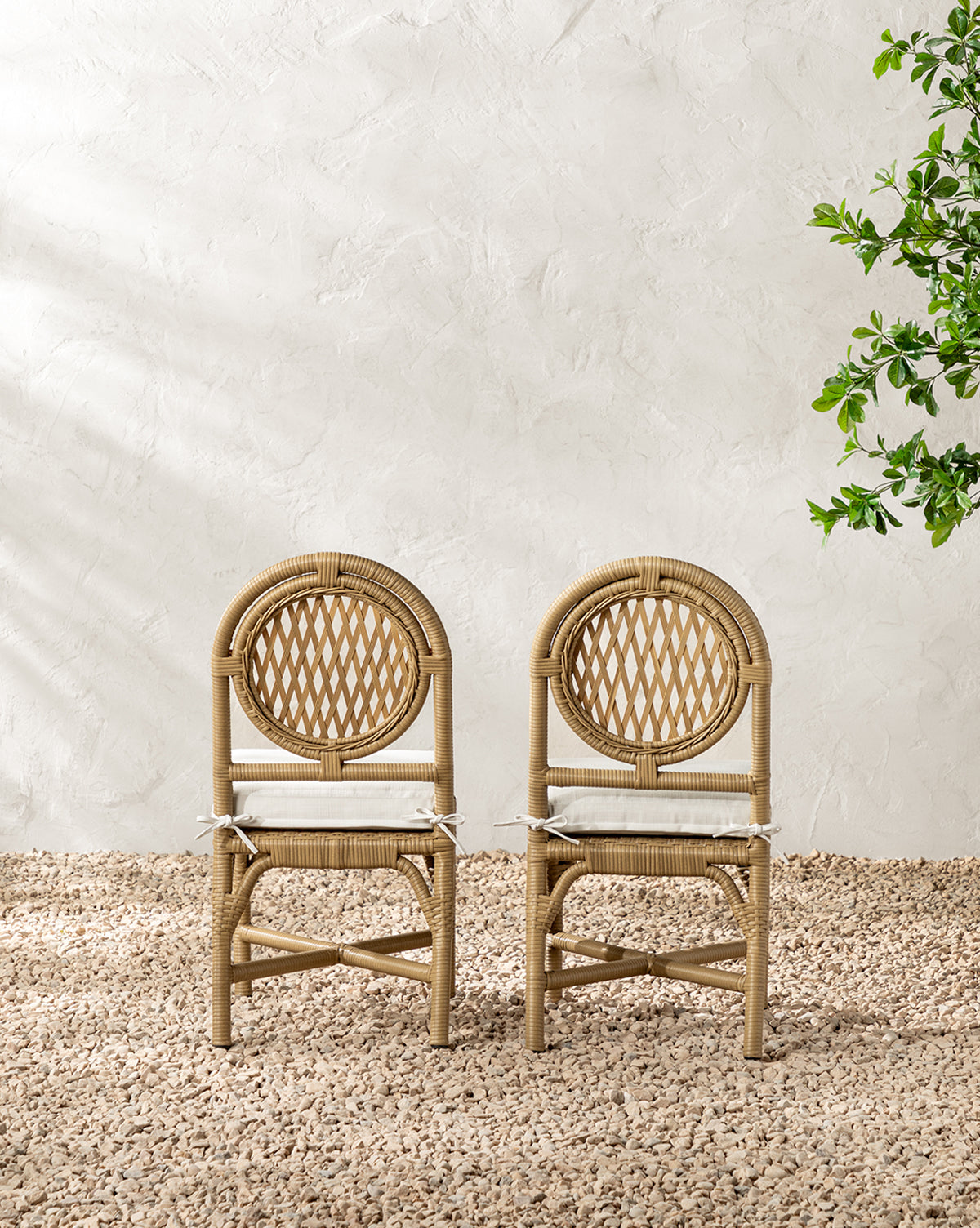 The Marisol Indoor/Outdoor Dining Chair (Set of 2), featuring weather-resistant woven rattan backs and seats, sits on a gravel floor before a white textured wall, with a leafy green plant to the right by McGee & Co.