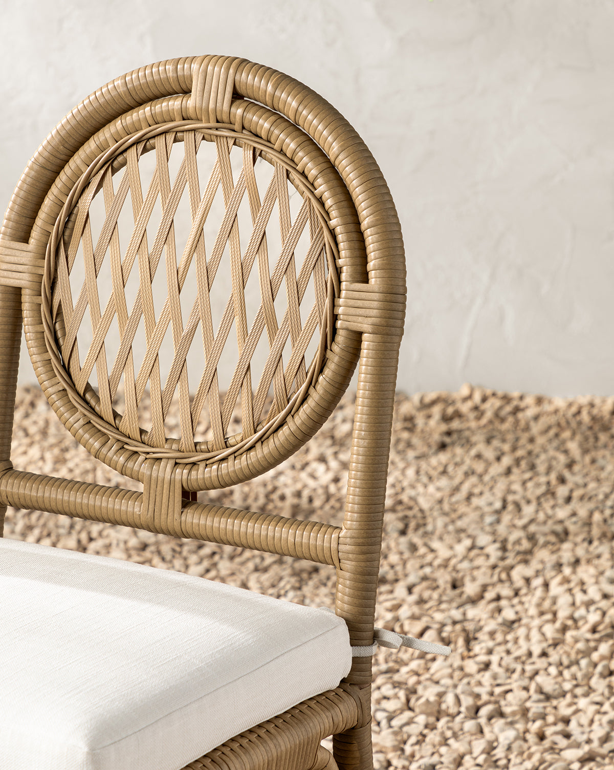 A close-up of the Marisol Indoor/Outdoor Dining Chair (Set of 2), featuring a weather-resistant, round handwoven wicker backrest and white cushioned seat, set on pebbles against a textured white wall, by McGee & Co.