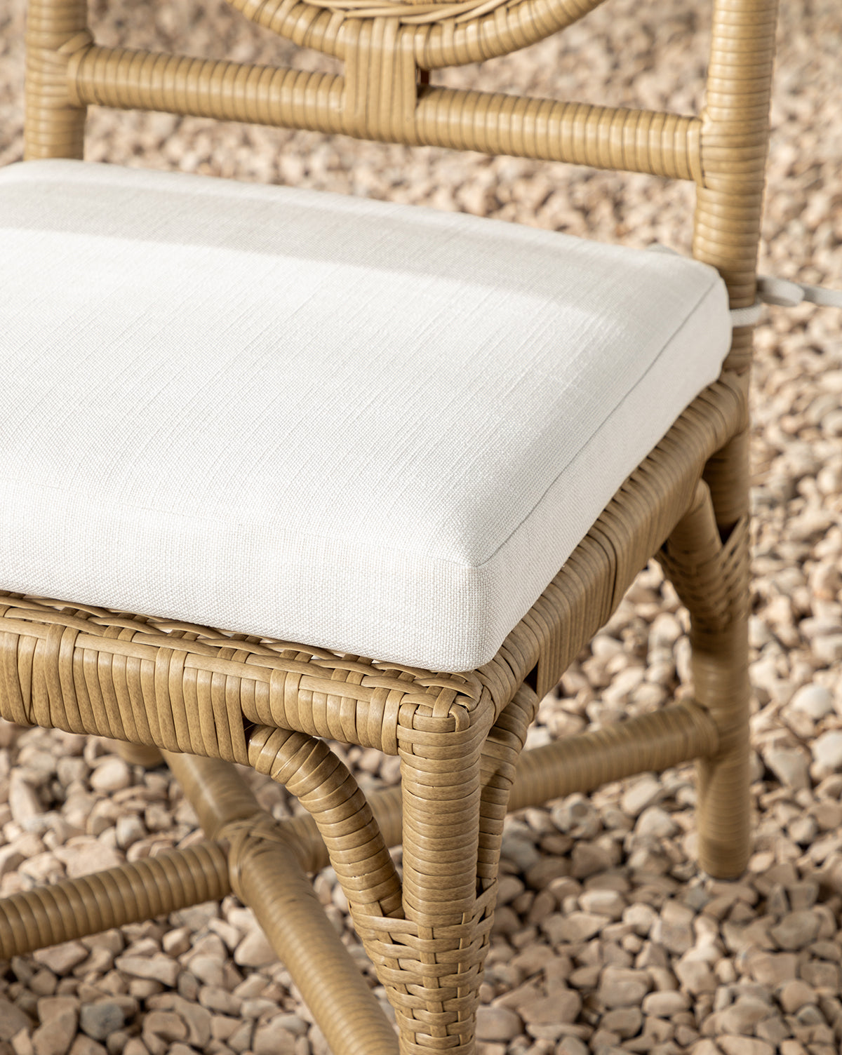 A close-up of the Marisol Indoor/Outdoor Dining Chair (Set of 2) featuring handwoven wicker and a white cushioned seat, placed outside on small beige pebbles, by McGee & Co.