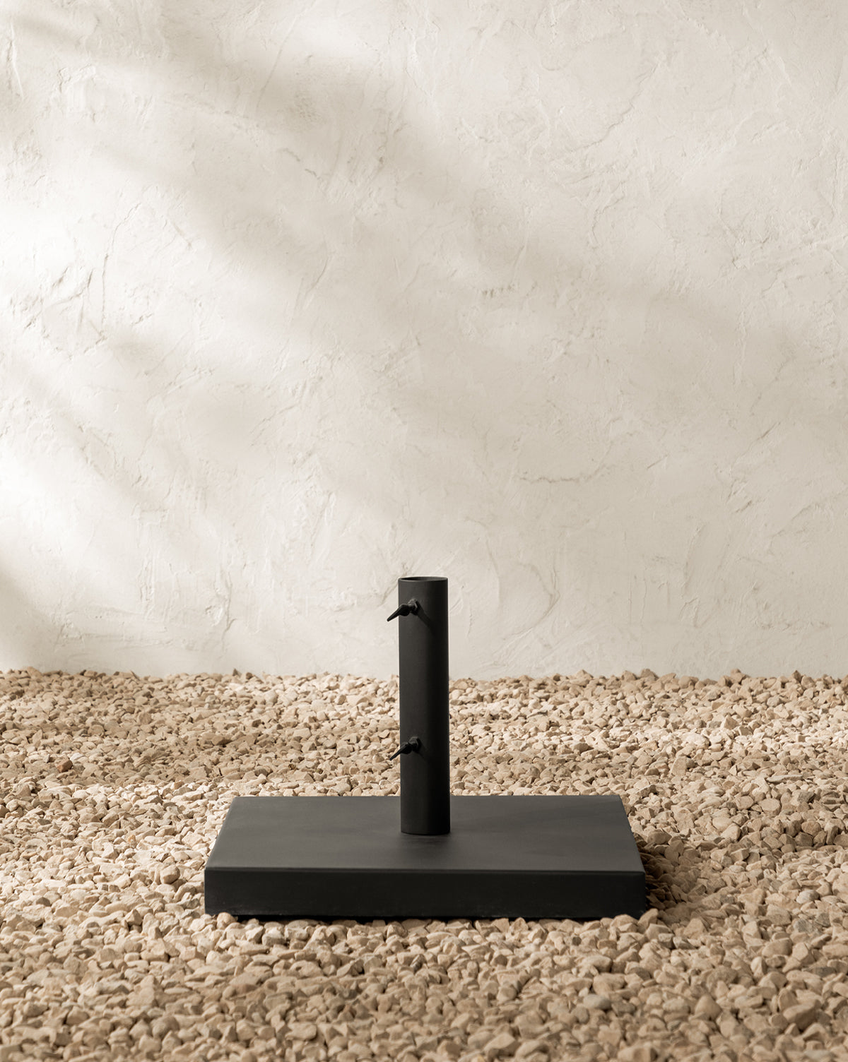 The Marques Outdoor Umbrella Stand, in a black finish with two adjustment knobs on its vertical pole, sits atop light-colored pebbles against a textured off-white wall, by McGee & Co.