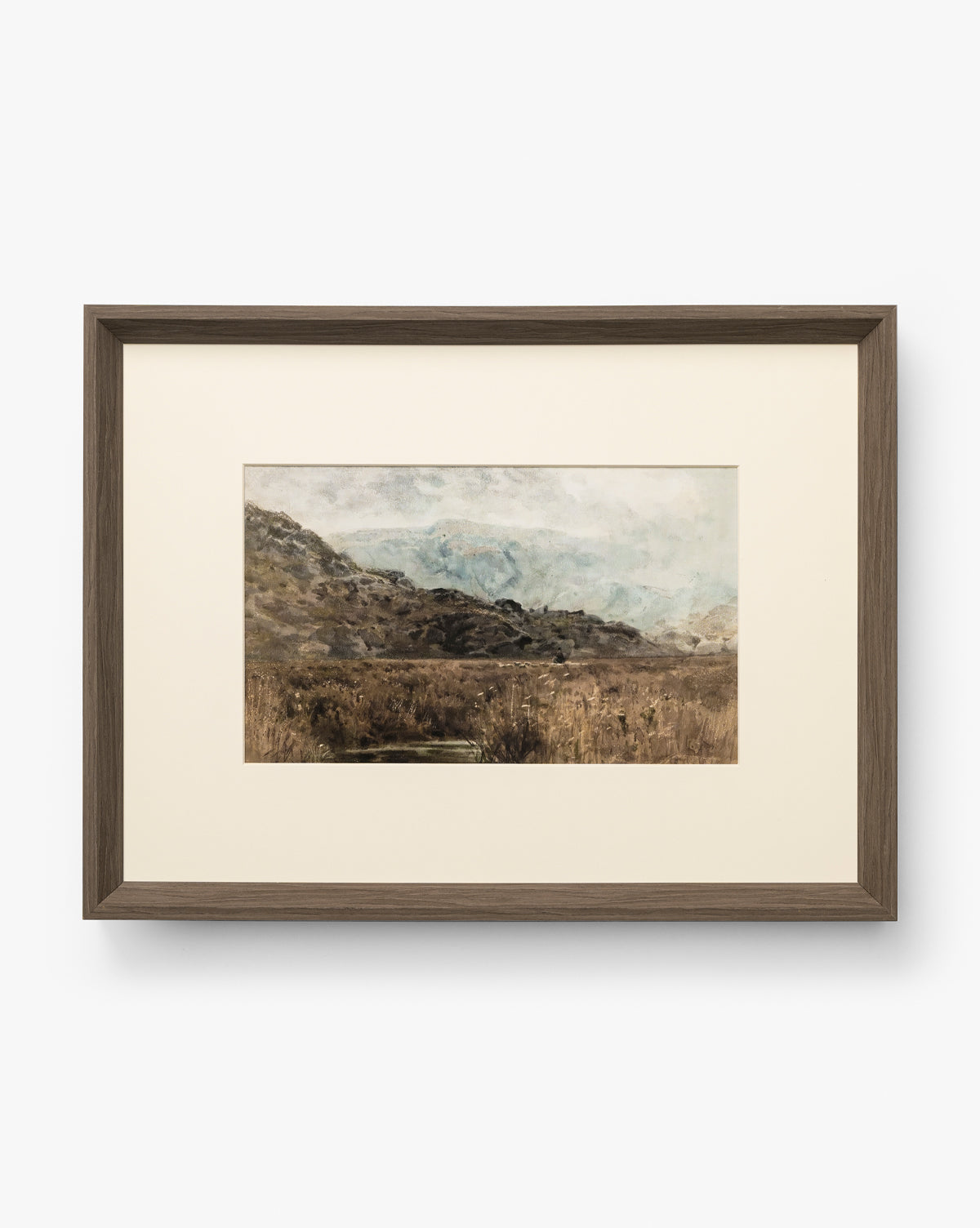 Marshland is a framed landscape painting with muted earthy tones, showing grassy hills and marshland beneath a cloudy sky. It features a simple, modern walnut woodgrain frame with a white mat for a soft, natural look by McGee & Co.