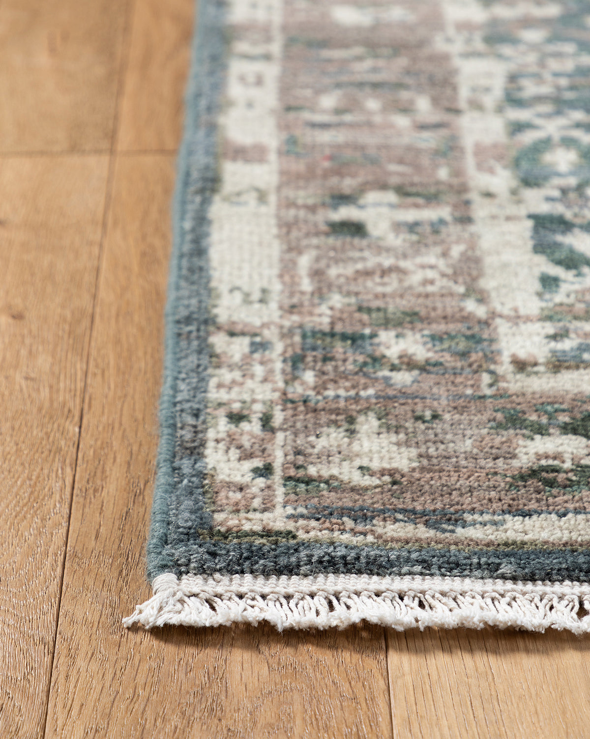 A close-up of the corner of the Marta Hand-Knotted Wool Rug, featuring blue, beige, and brown patterns with short white fringe, shown on a light wooden floor, by McGee & Co.