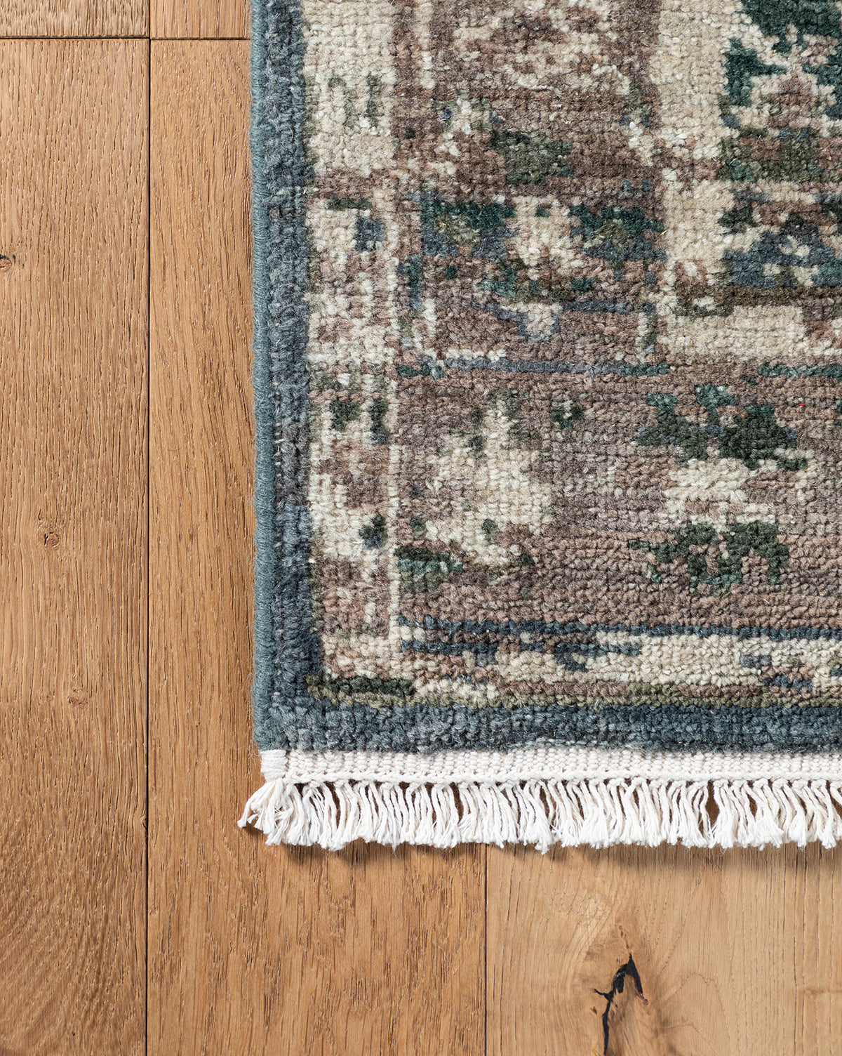 A close-up of the Marta Hand-Knotted Wool Rug with fringed edges on a wooden floor, showcasing its muted green, beige, and brown tones and worn vintage look, by McGee & Co.