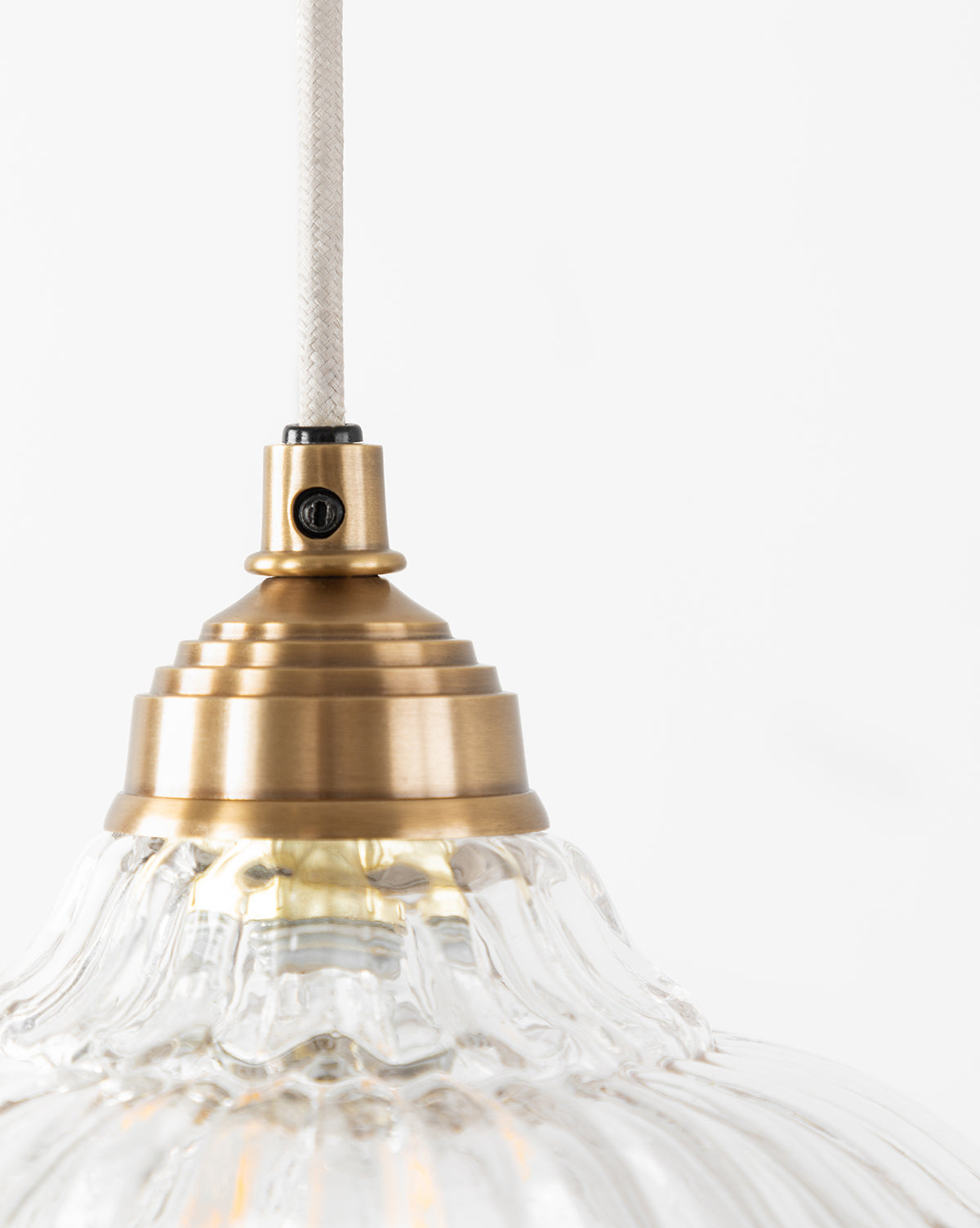 A close-up of the McGee & Co. x Loeffler Randall Maude Glass Pendant shows its ribbed clear glass shade and brass hardware, suspended by a white cord against a plain white background, by McGee & Co.