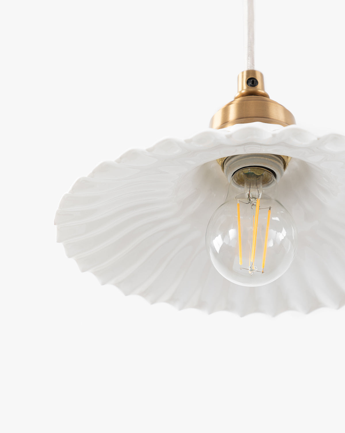 A close-up of the McGee & Co. x Loeffler Randall Maude Glass Pendant shows brass hardware, a clear round bulb, and a white wavy-edged ceramic shade, set against a plain white background by McGee & Co.