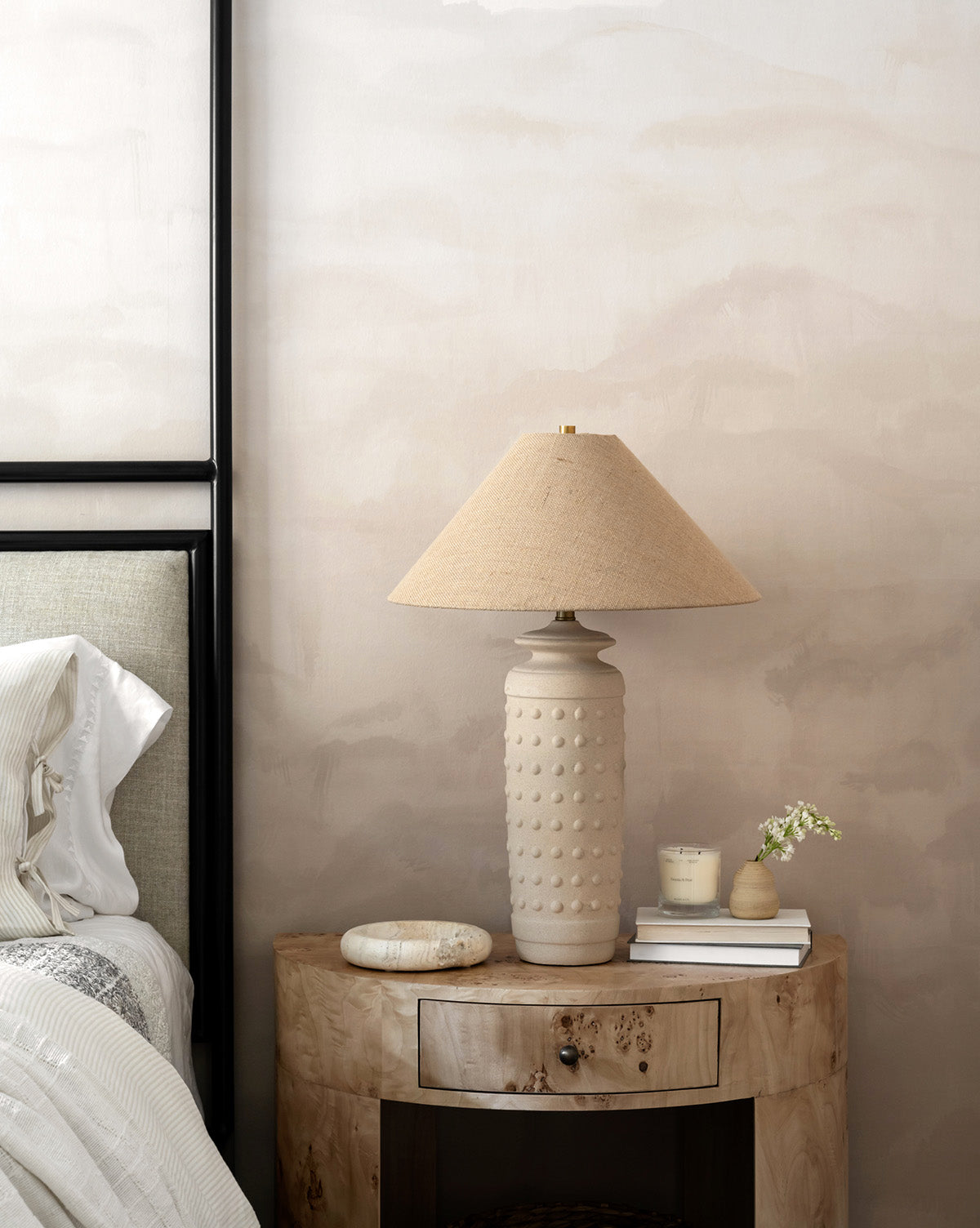 A neutral-toned bedroom corner features a wooden nightstand with a drawer, topped with the Sonoran Table Lamp, a candle, a decorative bowl, and a small vase with white flowers beside a bed with striped bedding, by McGee & Co.