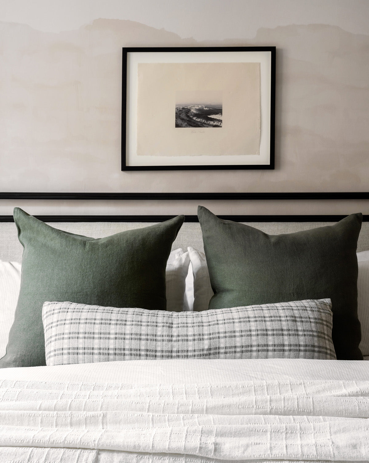 A neatly made bed with two large green pillows, a long plaid pillow, and white bedding sits beneath Palos Verdes, a museum-quality black and white ocean photograph, displayed on a muted, light-colored wall.