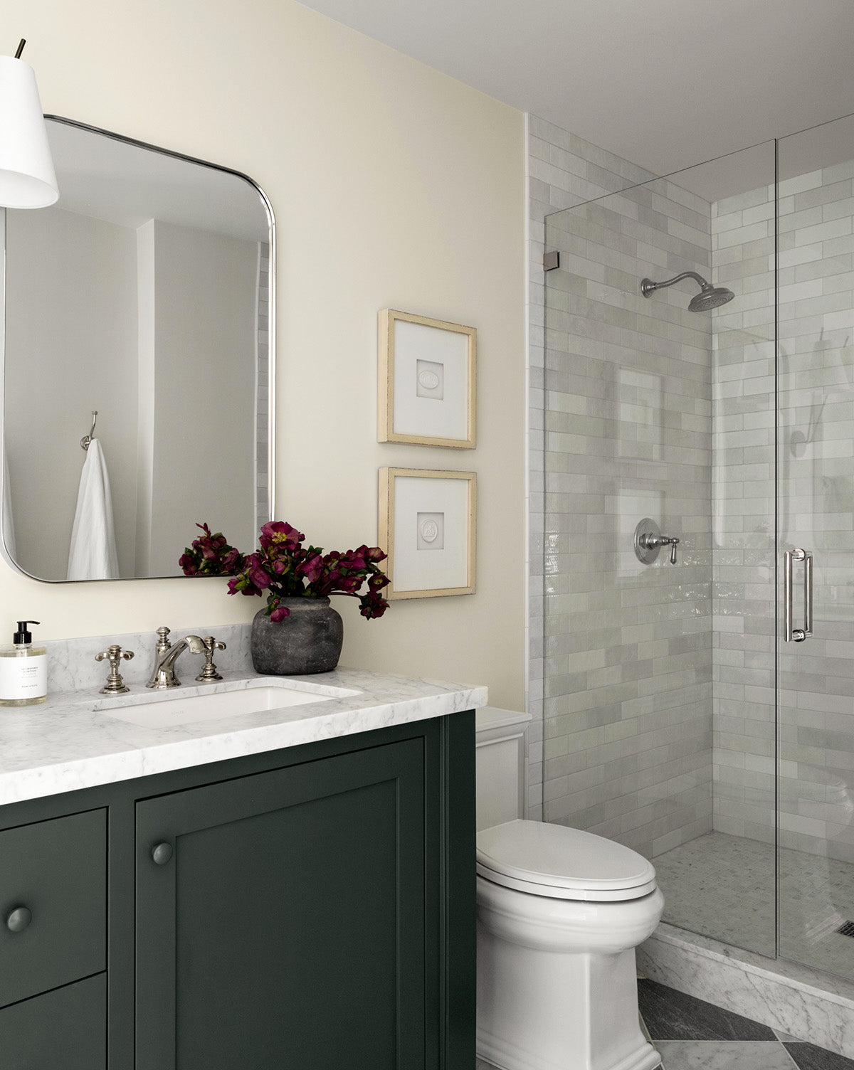 A modern bathroom features a dark green vanity, white countertop, silver fixtures, and Framed Intaglio II wall art beside the glass-enclosed shower with light tiles. A mirror and a vase of dark flowers sit above the sink.
