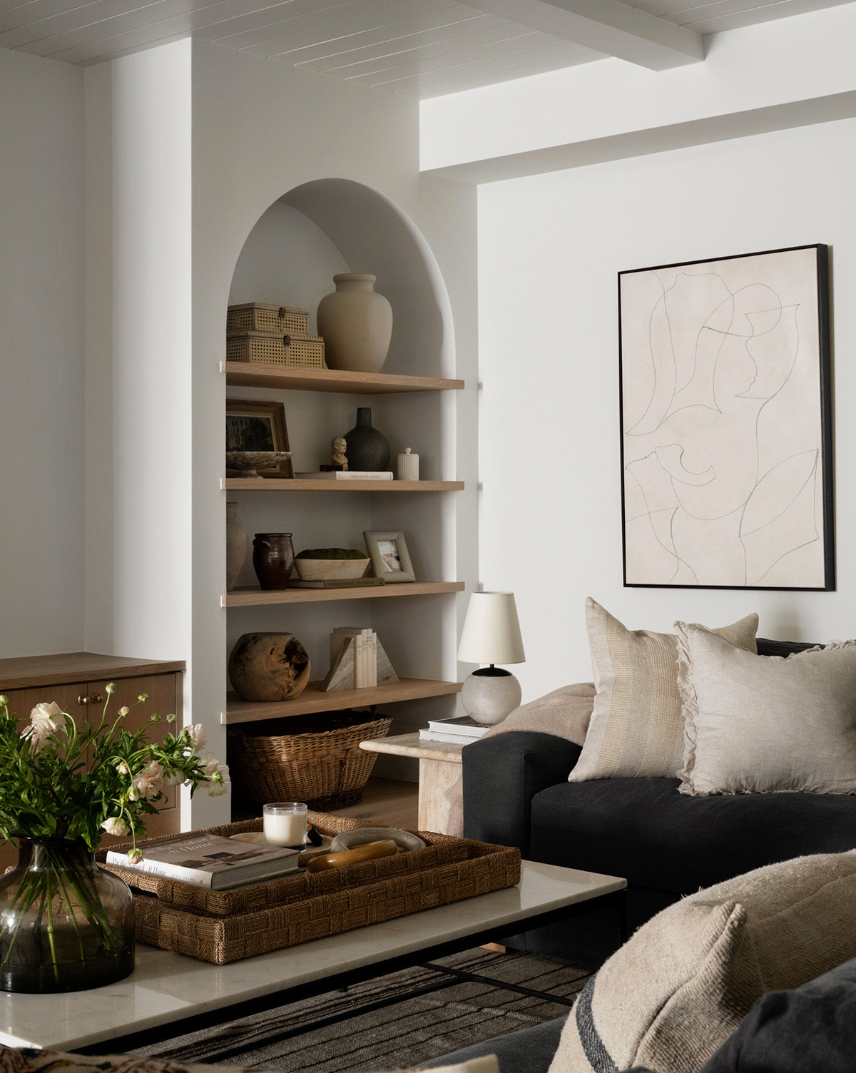 A cozy living room with neutral tones features a dark sofa, light cushions, a white coffee table with a wicker tray and vase, a built-in arched shelf with decor, and the Sketched Thoughts canvas art above the sofa.