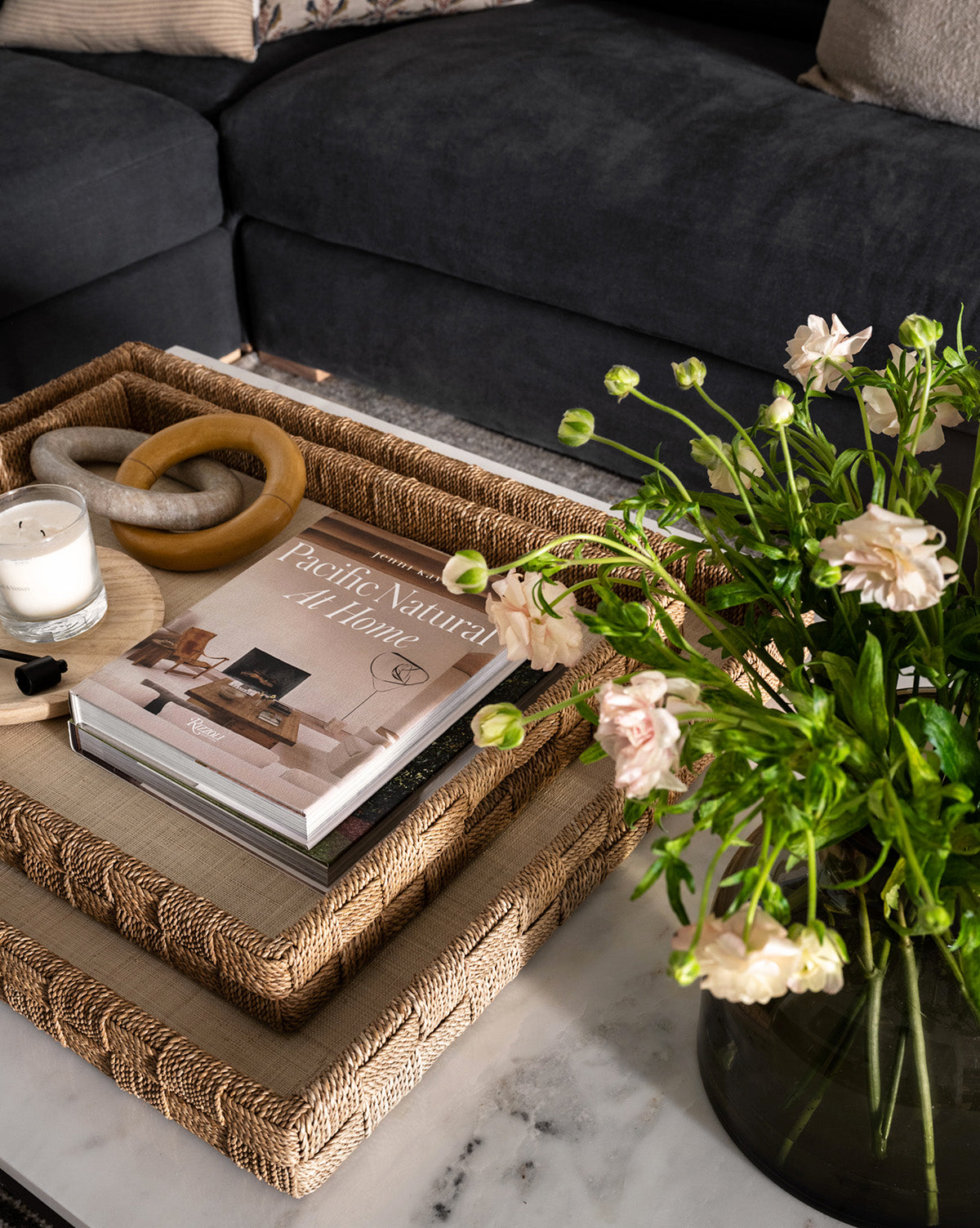 A cozy living room with a dark sofa, stacked woven trays on a marble coffee table, pale flowers in a vase, a candle, decorative objects, and the book Pacific Natural at Home—ideal for lovers of west coast-inspired interiors.