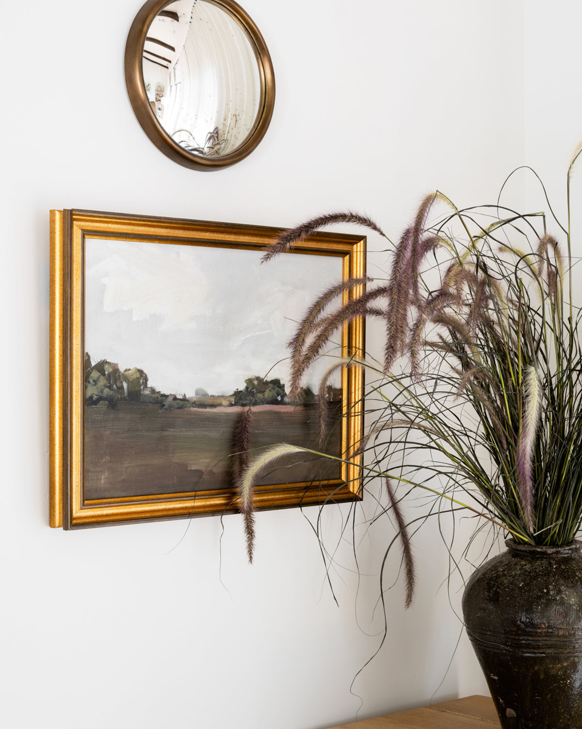 A round mirror and Meadow Landscape by Monique Lazard hang on a white wall beside a tall ceramic vase with wispy grass, creating an inviting home decor vignette.
