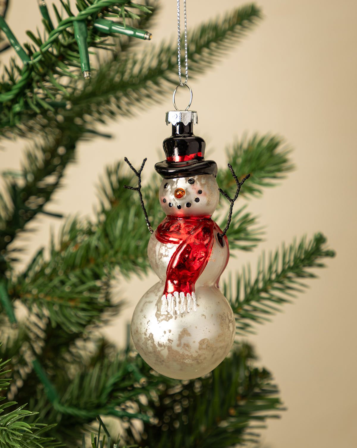 The Mercury Glass & Wire Snowman Ornament, featuring a red scarf, black top hat, and twig arms, hangs from a green Christmas tree branch, bringing vintage charm to your holiday décor against a warm beige background, by McGee & Co.