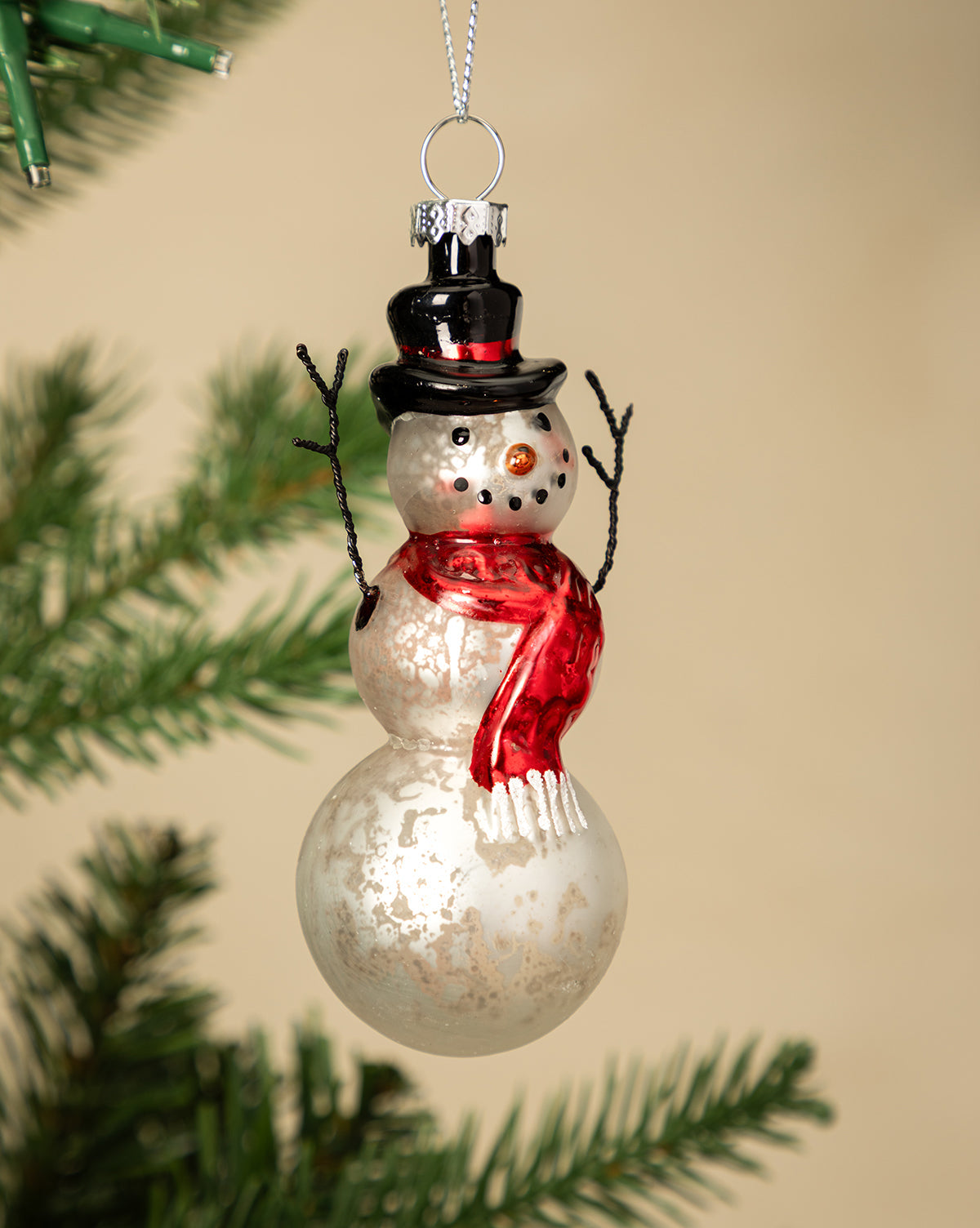 The Mercury Glass & Wire Snowman Ornament, featuring a black top hat, red scarf, and stick arms, hangs from a Christmas tree branch against a beige background, bringing vintage-inspired charm to your holiday decor, by McGee & Co.