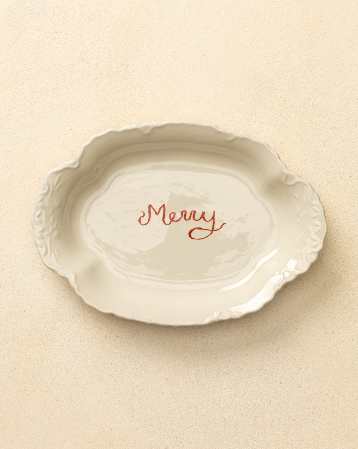The Merry Stoneware Scalloped Platter is an oval white platter with decorative edges and "Merry" in red script at the center, perfect for holiday serving and festive table decor, by McGee & Co.