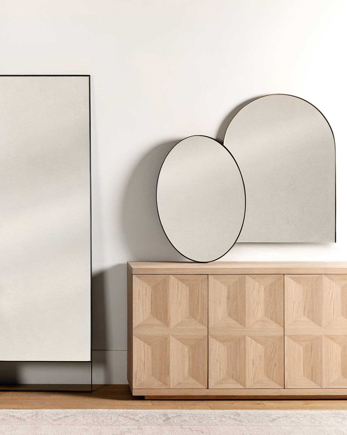 Minimalist room featuring a light wood geometric sideboard topped with the Hogan Oval Mirror in an elegant stainless steel frame, an arched mirror leaning above, and a large rectangular mirror standing beside the sideboard, by McGee & Co.