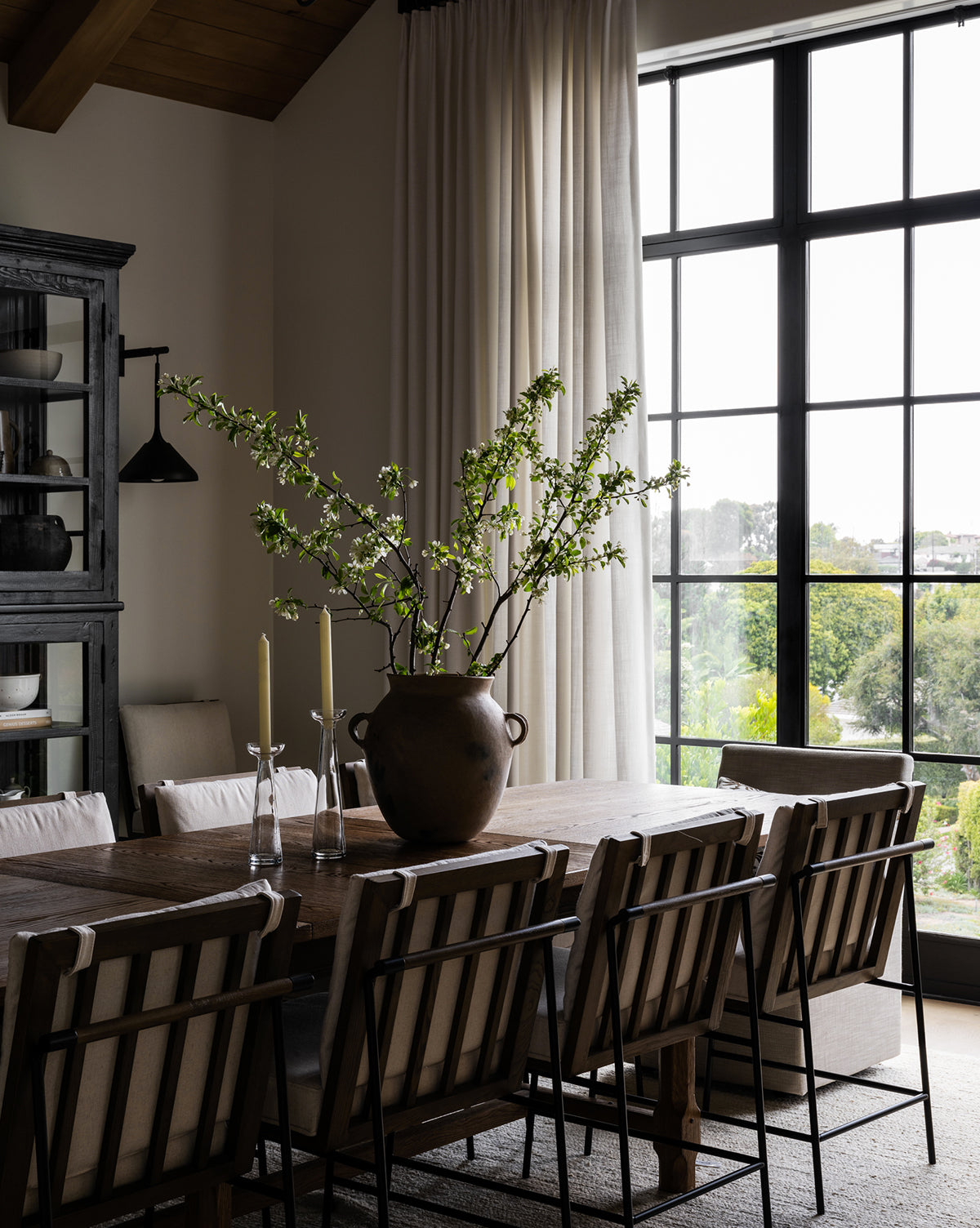 A dining room with a wooden table and eight Selas Dining Chairs with black iron frames. Tall white curtains dress the large window. On the table, there’s a vase of green branches and two candlesticks. Natural light enhances the neutral decor, by McGee & Co.