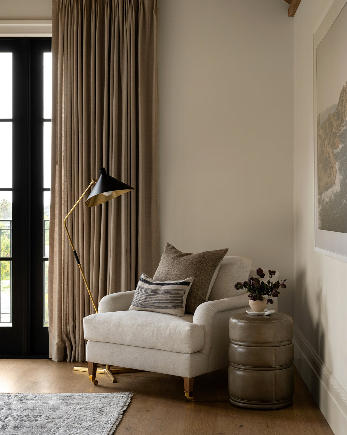 A cozy living room corner features a white armchair with neutral pillows, the Mayotte Offset Floor Lamp in antique brass with a black shade, beige curtains, a round side table, a potted plant, wall art, and light wood flooring by McGee & Co.