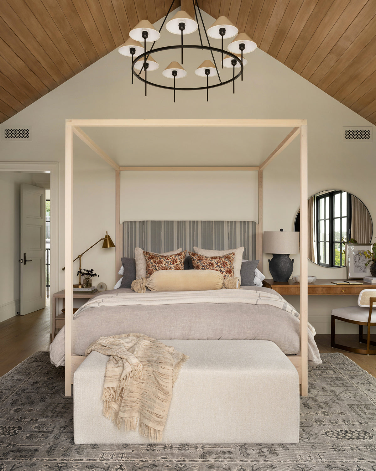 A modern bedroom features a four-poster bed with neutral bedding, decorative pillows, a bench and throw, patterned rug, the Anza Table Lamp in black ash glaze, and a vanity with round mirror under a vaulted wood ceiling and chandelier, by McGee & Co.